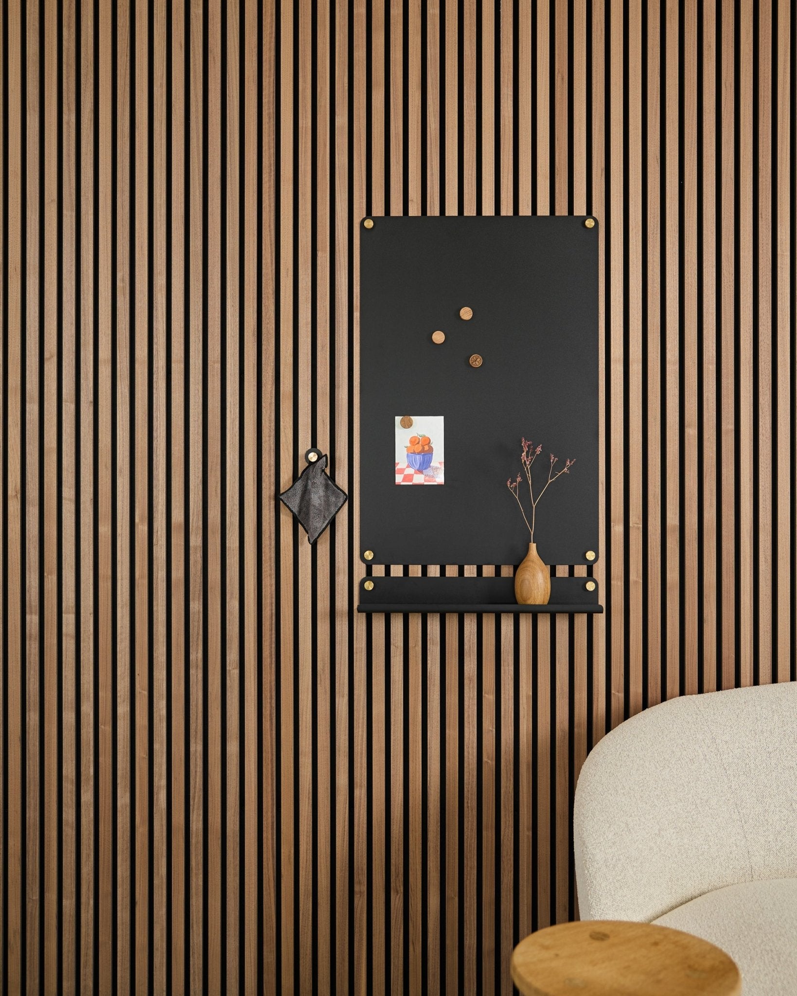 A large black Norr Magnetic Board by WoodUpp, designed for acoustic panels, hangs beside a white upholstered chair with notes, a vase, and a trinket.